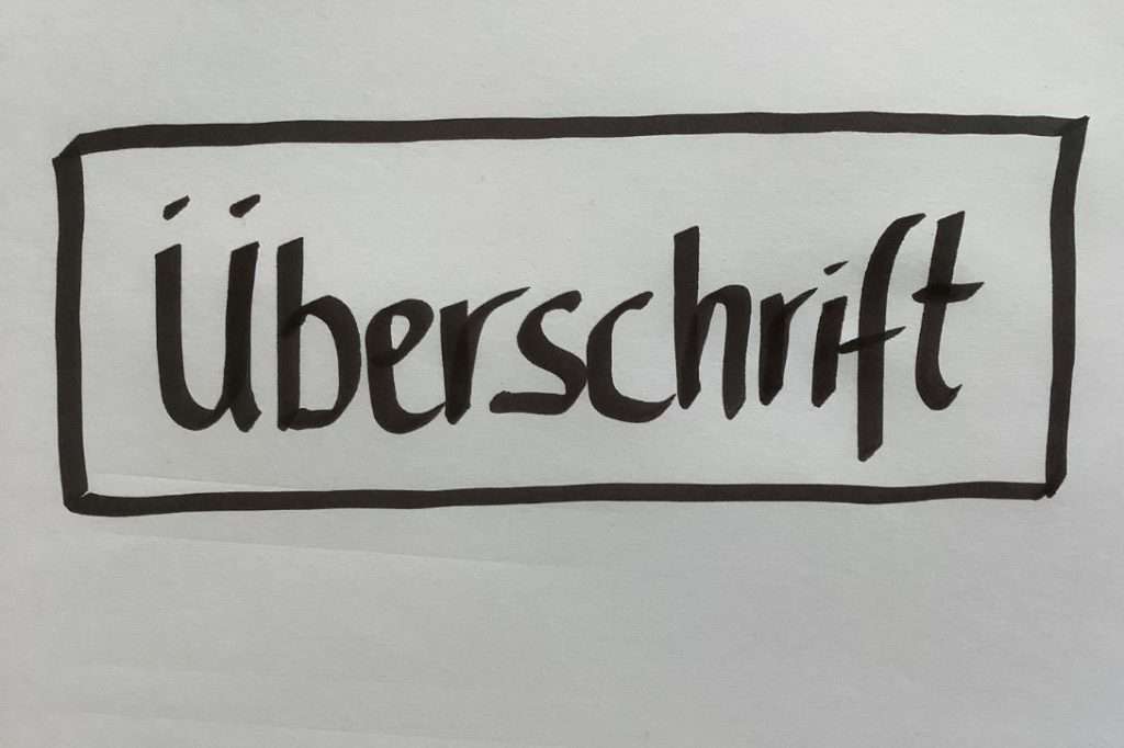 Auf dem Bild ist das Wort „Überschrift“ zu sehen. Es ist mit einem dicken, schwarzen Filzstift geschrieben. Um das Wort herum ist ein schwarzer Kasten gemalt. Der Rahmen hebt das Wort gut sichtbar hervor. Die Buchstaben sind groß und klar lesbar. Diese Gestaltung hilft dabei, den Blick sofort auf das Thema oder den Titel zu lenken. Das Bild zeigt: Wenn Du Flipcharts gestalten willst, kannst Du eine Überschrift mit Rahmen hervorheben, damit sie nicht übersehen wird.