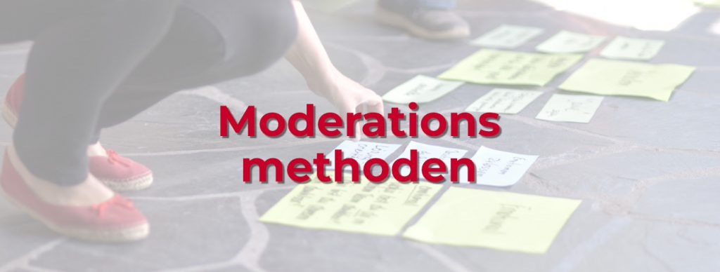 Person nimmt an einem Workshop teil und arbeitet mit Moderationsmethoden. Sie kniet auf einem dunklen Steinboden und zeigt auf eine Karte mit handgeschriebenem Text. Vor ihr liegen zahlreiche bunte Moderationskarten, vorwiegend in Grün und Weiß, die verschiedene Stichpunkte enthalten. Eine zweite Person steht im Hintergrund, erkennbar an den Schuhen. In der linken Hand hält die kniende Person ein Heft oder einen Block.
