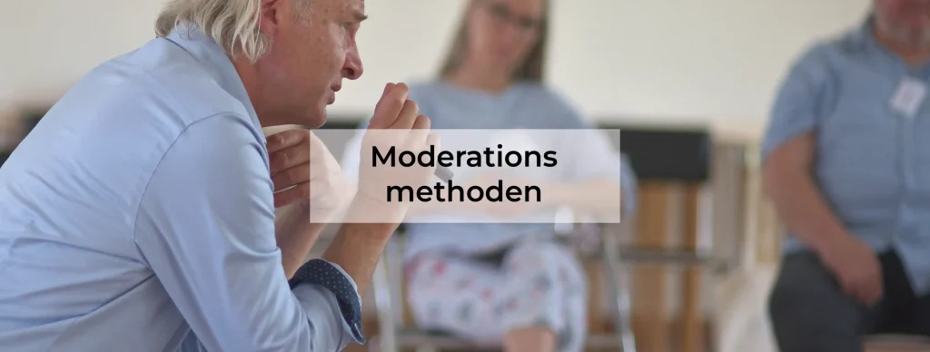 Person nimmt an einem Workshop teil und arbeitet mit Moderationsmethoden. Sie kniet auf einem dunklen Steinboden und zeigt auf eine Karte mit handgeschriebenem Text. Vor ihr liegen zahlreiche bunte Moderationskarten, vorwiegend in Grün und Weiß, die verschiedene Stichpunkte enthalten. Eine zweite Person steht im Hintergrund, erkennbar an den Schuhen. In der linken Hand hält die kniende Person ein Heft oder einen Block.