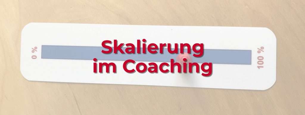 Das Bild zeigt eine Skala zur Skalierung im Coaching, die auf einem Holztisch liegt. Die Skala ist eine rechteckige Karte mit abgerundeten Ecken. Sie hat eine lange, horizontale Leiste in der Mitte, die als Messbereich dient. Links steht „0 %“, rechts „100 %“. Ein roter, runder Marker ist auf der Skala platziert, um eine bestimmte Position anzuzeigen. Darüber liegt ein roter Schriftzug mit dem Text „Skalierung im Coaching“, der leicht transparent wirkt. Diese Skalierung im Coaching wird genutzt, um subjektive Einschätzungen sichtbar zu machen. Coachees können auf einer Skala angeben, wie sie ihren aktuellen Zustand bewerten oder wie weit sie ein Ziel bereits erreicht haben.