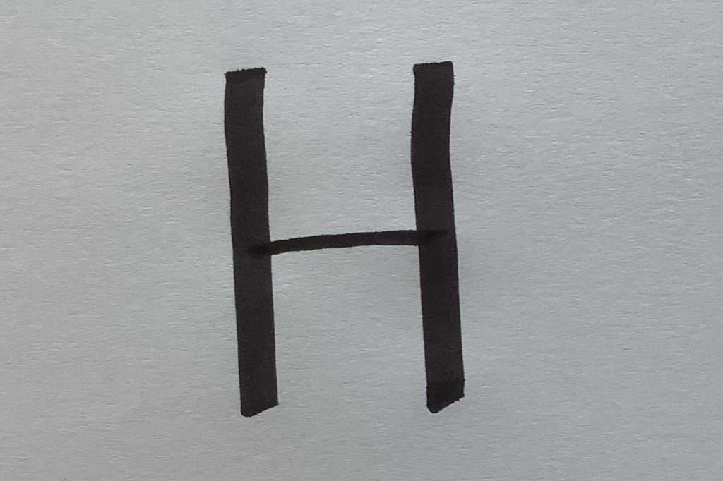 Das Bild zeigt den Buchstaben „H“, der mit einem schwarzen Filzstift auf weißem Papier geschrieben wurde. Die beiden senkrechten Striche links und rechts sind dick und gleichmäßig. Der waagerechte Mittelstrich ist dünner. Wenn Du Flipcharts gestalten willst, achte auf eine gute Stifthaltung. So wirken Deine Buchstaben klar, kräftig und gut lesbar – auch aus der Entfernung.