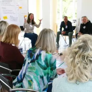 Auf dem Bild ist eine Seminarsituation zu sehen. Mehrere Teilnehmende sitzen im Kreis. Vorne sitzt die Trainerin, die gerade gestikuliert. Hinter der Trainerin steht eine Pinnwand.