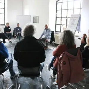 Das Bild zeigt einen Raum mit mehreren Teilnehmenden. Sie sitzen im Kreis. Vorne sitzt der Trainer. Neben dem Trainer steht ein Flipchart,