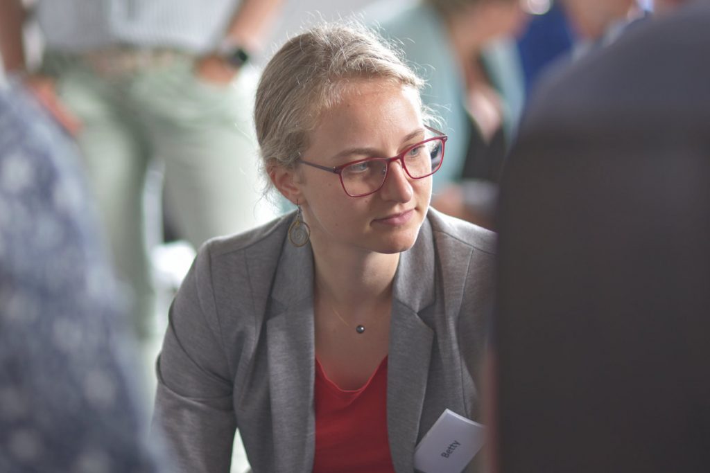 Auf dem Bild ist eine junge Frau mit blonden Haaren und roter Brille zu sehen. Sie trägt eine graue Jacke und ein rotes Oberteil. Sie schaut aufmerksam zur Seite, als ob sie jemandem zuhört. Im Hintergrund sind mehrere Menschen leicht verschwommen erkennbar – sie stehen oder sitzen in einer Gesprächsrunde. Die Stimmung wirkt ruhig und konzentriert.

Das Bild zeigt eine Szene aus der Basisausbildung Systemischer Business Coach. Es macht deutlich, wie persönlich und nahbar die Weiterbildung bei abb-seminare gestaltet ist. Die Teilnehmenden arbeiten im Austausch miteinander und lernen voneinander.