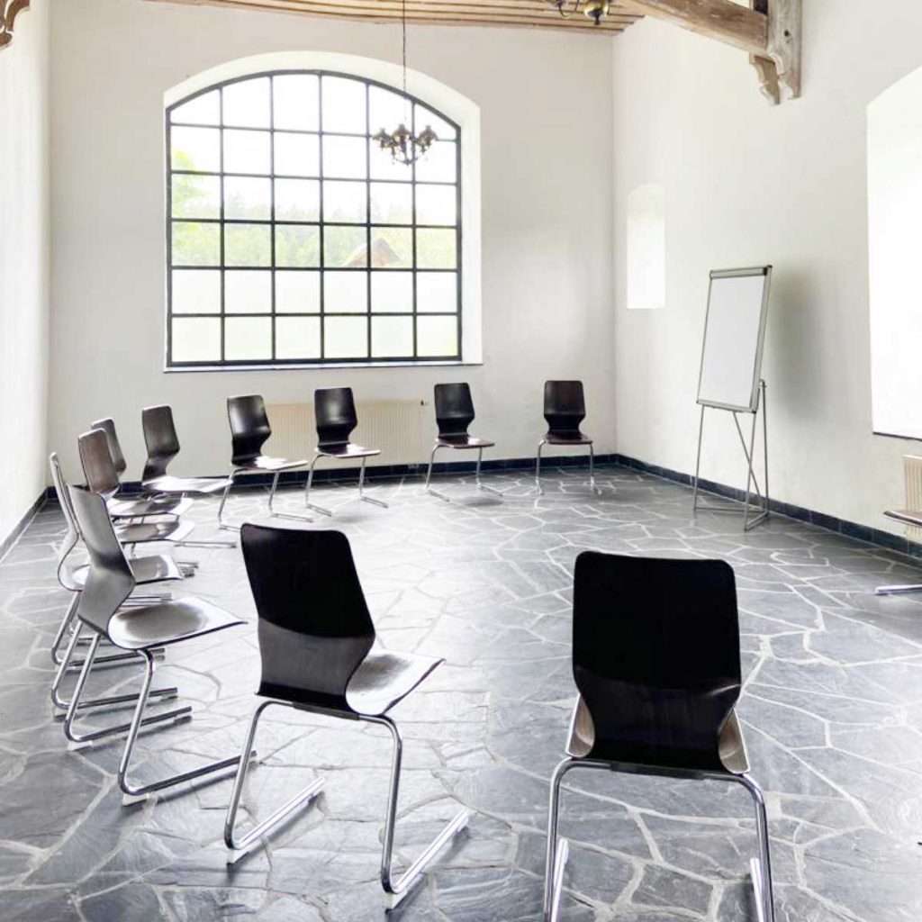 Das Bild zeigt einen hellen Seminarraum mit einem großen Fenster, durch das Tageslicht in den Raum fällt. Der Boden besteht aus grauen Schieferplatten, die den Raum besonders wirken lassen. Im Kreis stehen moderne schwarze Stühle mit Metallgestell. An der Wand rechts ist ein Flipchart aufgestellt. Die weißen Wände und die hohe Decke mit sichtbaren Holzbalken geben dem Raum eine offene und freundliche Atmosphäre. Dieser Raum wird bei den abb-Impuls-Tagen genutzt.