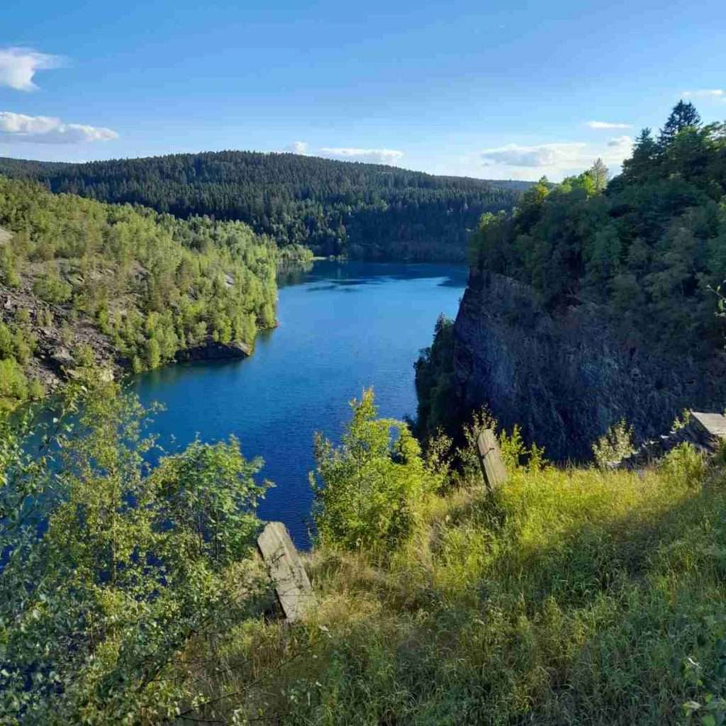 Das Bild zeigt einen tiefblauen See, der von grünen Bäumen und felsigen Klippen umgeben ist. Am Ufer wachsen Gräser und Sträucher, die ins Bild hineinragen. Im Hintergrund erstreckt sich ein dichter Wald bis zum Horizont. Der Himmel ist klar und blau, nur wenige weiße Wolken sind zu sehen. Die Landschaft wirkt ruhig und einladend und lädt zu Naturerlebnissen ein. Diese Umgebung gehört zum Veranstaltungsort der abb-Impuls-Tage.