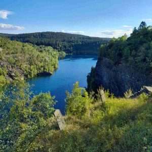 Das Bild zeigt einen tiefblauen See, der von grünen Bäumen und felsigen Klippen umgeben ist. Am Ufer wachsen Gräser und Sträucher, die ins Bild hineinragen. Im Hintergrund erstreckt sich ein dichter Wald bis zum Horizont. Der Himmel ist klar und blau, nur wenige weiße Wolken sind zu sehen. Die Landschaft wirkt ruhig und einladend und lädt zu Naturerlebnissen ein. Diese Umgebung gehört zum Veranstaltungsort der abb-Impuls-Tage.