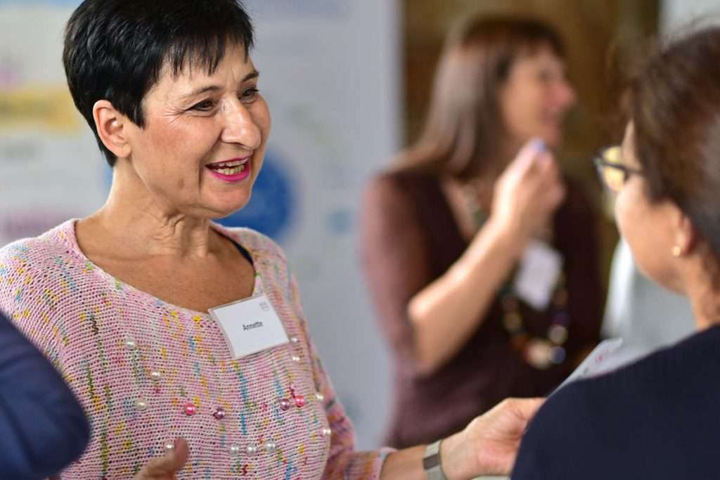 Eine Frau mit kurzem dunklem Haar spricht lächelnd mit einer anderen Teilnehmerin. Sie trägt einen bunten Pullover in Rosa mit kleinen Perlen. Im Hintergrund unterhalten sich weitere Menschen. Das Bild zeigt eine offene und freundliche Lernatmosphäre, in der Menschen miteinander ins Gespräch kommen. Es passt zum Thema Ausbildung Systemischer Business Coach – Coaching Grundkenntnisse erwerben, weil hier persönlicher Austausch und erste Übungen im Mittelpunkt stehen.