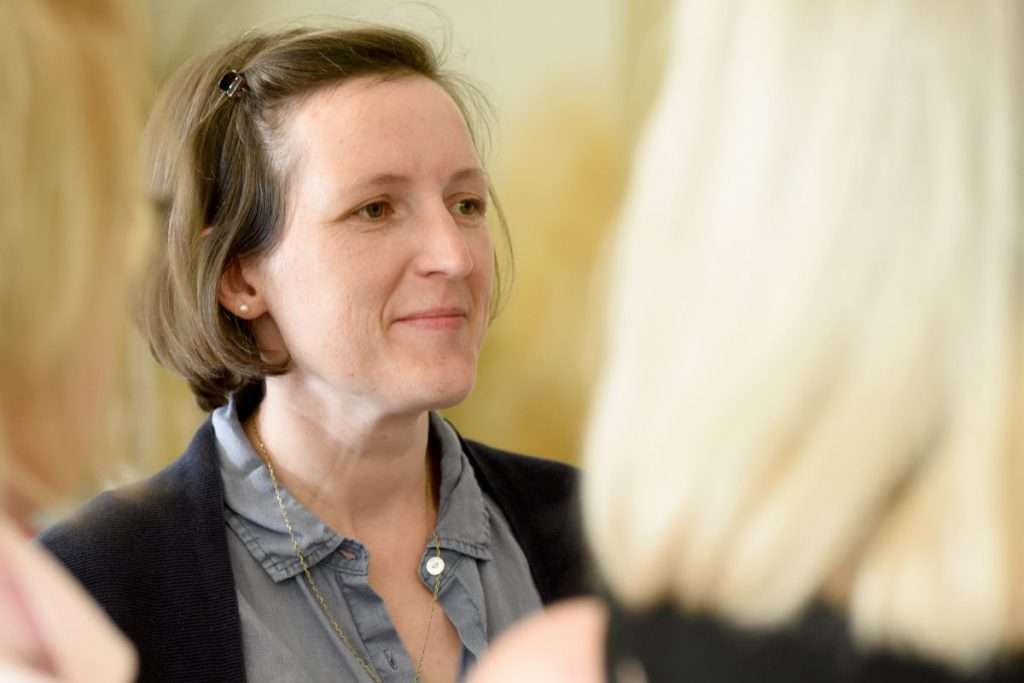 Eine Frau mit kurzen braunen Haaren und einer dunklen Jacke schaut aufmerksam nach vorn. Vor ihr ist unscharf der Hinterkopf einer anderen Person mit blonden Haaren zu sehen. Es passt zum Thema Weiterbildung Coaching – Coaching Kenntnisse vertiefen und zeigt, wie wichtig achtsames Zuhören im Coaching ist. Das Bild zeigt, wie wichtig persönlicher Austausch und Vernetzung in einer Coaching Ausbildung – Coachingprofil schärfen sind.