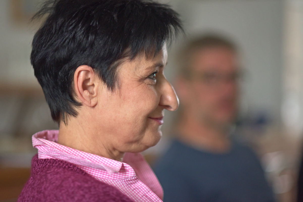 Das Foto zeigt eine Frau im Profil von der Seite. Sie hat kurze dunkle Haare und trägt ein pinkes Oberteil mit einem hellen Kragen. Ihr Gesicht wirkt ruhig und aufmerksam. Sie lächelt leicht. Im Hintergrund sind andere Menschen unscharf zu sehen. Die Frau hört konzentriert zu und scheint an einer Übung oder einem Gespräch teilzunehmen. Das Bild passt gut zur NLP Basic Ausbildung, weil es Lernen, Zuhören und persönliche Entwicklung in einer ruhigen und wertschätzenden Lernatmosphäre zeigt.