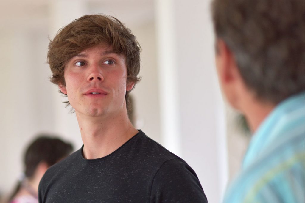 Das Foto zeigt einen jungen Mann mit lockigen braunen Haaren. Er trägt ein dunkles T-Shirt. Er steht in einem hellen Raum und schaut aufmerksam zu einer anderen Person, die rechts im Bild unscharf zu sehen ist. Sein Gesicht wirkt offen und interessiert. Im Hintergrund sind weitere Menschen verschwommen zu erkennen. Die Szene zeigt einen ruhigen Austausch und aktives Zuhören. Das Bild passt zur NLP Practitioner Ausbildung, weil es Lernen, Kommunikation und persönliche Entwicklung in einer Gruppe zeigt.
