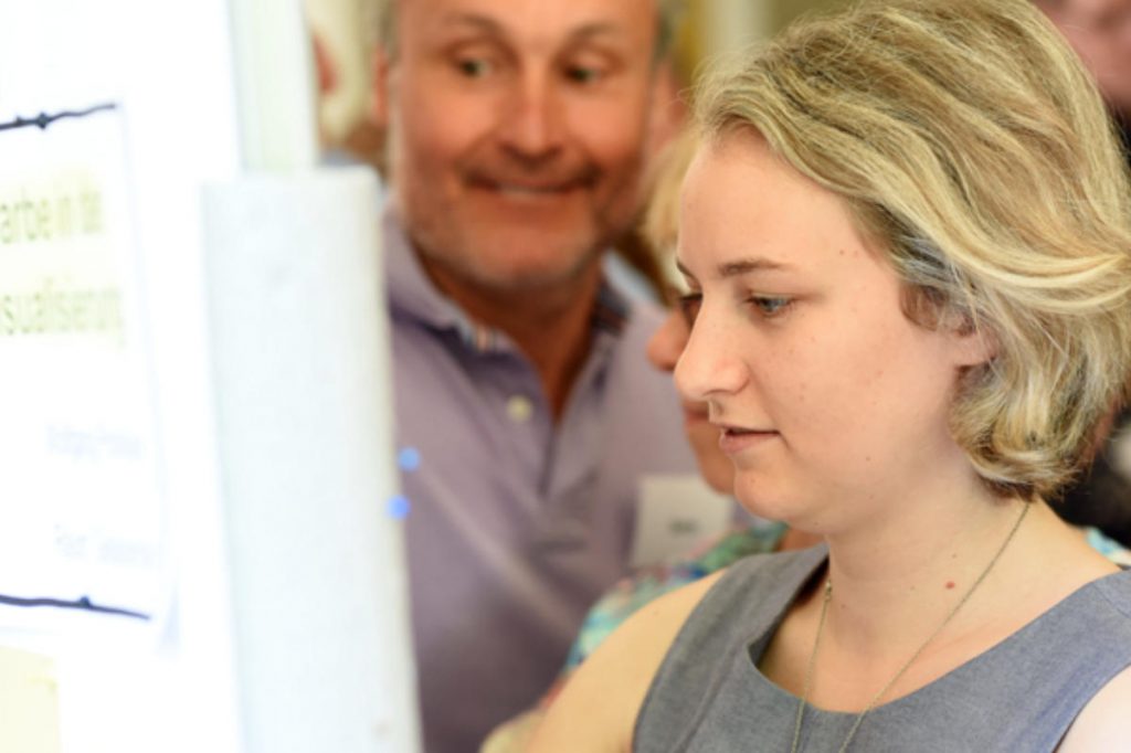 Eine junge Frau steht dicht vor einer Pinnwand und liest konzentriert einen Zettel. Hinter ihr stehen weitere Personen, die interessiert zuschauen. Das Bild zeigt eine aktive Lernsituation, wie sie in einer Trainerausbildung modular typisch ist. Die Menschen arbeiten gemeinsam an einer Aufgabe und tauschen sich aus.