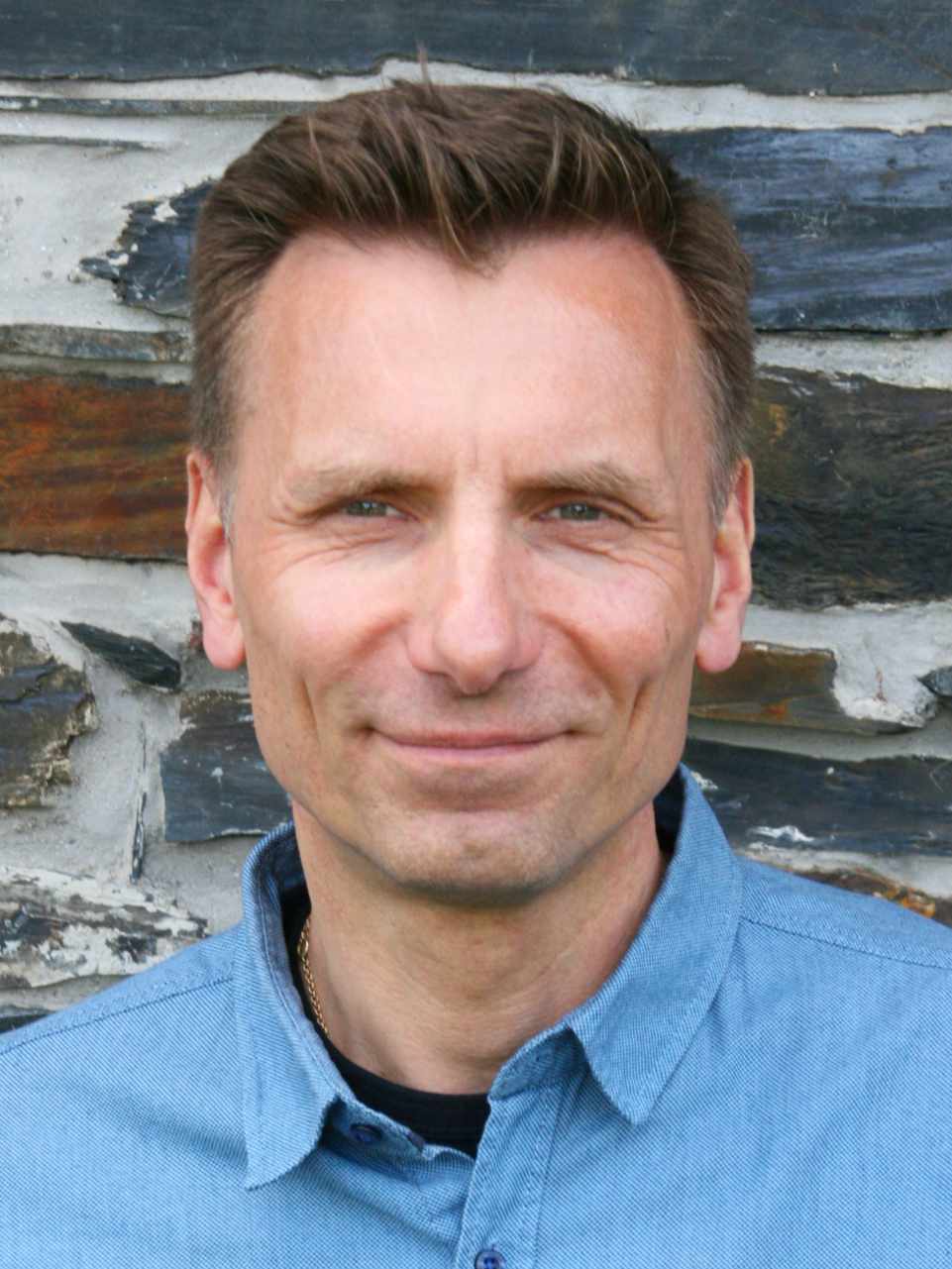 Du siehst ein Porträt von einer erwachsenen Person. Die Person schaut direkt in die Kamera. Das Gesicht ist ruhig und freundlich. Die Person lächelt leicht. Die Haare sind kurz und braun. Die Person trägt ein hellblaues Hemd. Der Hintergrund besteht aus einer Steinwand mit grauen und dunklen Steinen. Das Bild zeigt Ausbildungsleiter René Penselin der NLP Ausbildung.