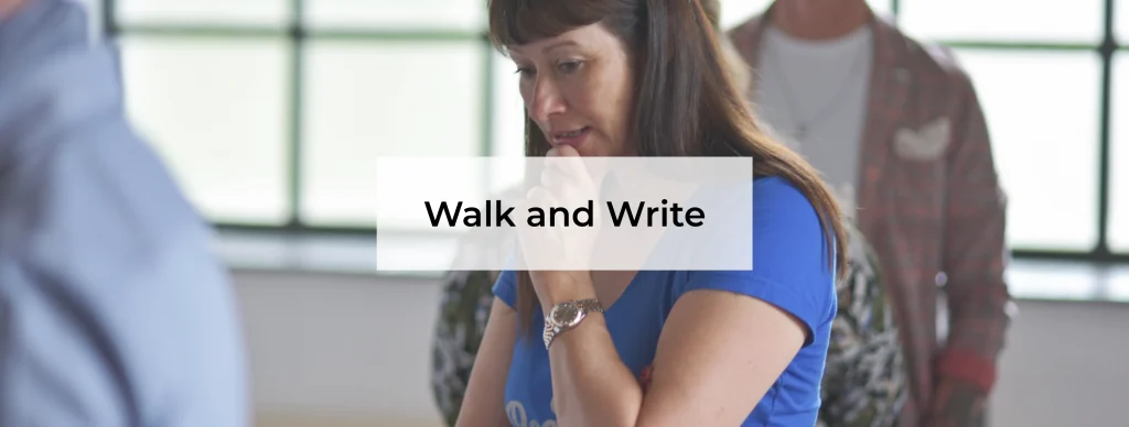 Eine Frau steht in einem hellen Seminarraum und denkt nach. Sie trägt ein blaues T-Shirt und hält ihre Hand an das Kinn. Ihr Blick ist nach unten gerichtet, als würde sie sich auf eine Aufgabe konzentrieren. Im Hintergrund stehen weitere Personen, leicht unscharf. Die Szene zeigt eine ruhige Arbeitsphase in einer Gruppe. Die Moderationstechnik Walk & Write wird genutzt, um Gedanken zu sammeln, Ideen zu ordnen und gemeinsam Lösungen zu entwickeln.