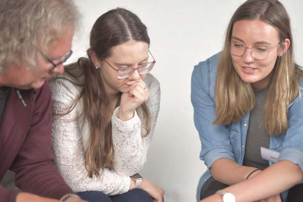 Das Bild zeigt drei Menschen, die gemeinsam an einer Aufgabe arbeiten. Zwei junge Frauen und ein älterer Mann beugen sich über einen Tisch und schauen konzentriert auf etwas, das außerhalb des Bildes liegt. Eine der Frauen hält nachdenklich ihre Hand an das Kinn, während die andere spricht und ihre Gedanken einbringt. Die Gruppe wirkt aufmerksam, interessiert und engagiert. Das Motiv passt gut zum Thema NLP Basic Ausbildung - Klar kommunizieren & herausfordernde Situationen souverän meistern. Es zeigt Menschen, die ihre Kommunikationsfähigkeiten verbessern, unterschiedliche Sichtweisen verstehen und gemeinsam neue Wege für den Umgang mit beruflichen und persönlichen Herausforderungen entwickeln.
