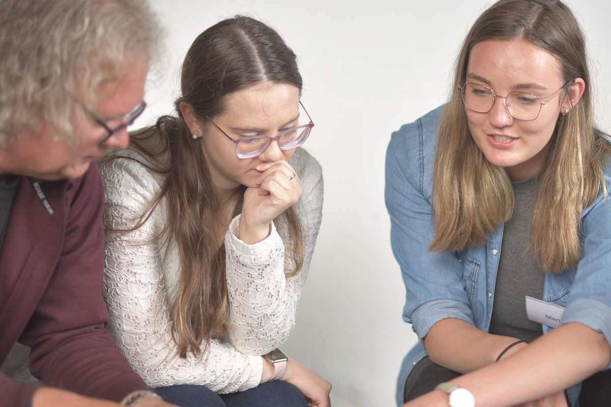 Das Bild zeigt drei Menschen, die gemeinsam an einer Aufgabe arbeiten. Zwei junge Frauen und ein älterer Mann beugen sich über einen Tisch und schauen konzentriert auf etwas, das außerhalb des Bildes liegt. Eine der Frauen hält nachdenklich ihre Hand an das Kinn, während die andere spricht und ihre Gedanken einbringt. Die Gruppe wirkt aufmerksam, interessiert und engagiert. Das Motiv passt gut zum Thema NLP Basic Ausbildung - Klar kommunizieren & herausfordernde Situationen souverän meistern. Es zeigt Menschen, die ihre Kommunikationsfähigkeiten verbessern, unterschiedliche Sichtweisen verstehen und gemeinsam neue Wege für den Umgang mit beruflichen und persönlichen Herausforderungen entwickeln.