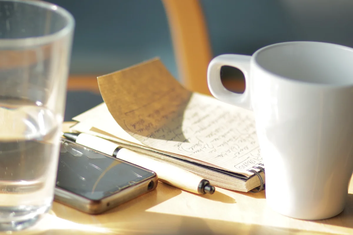 Das Bild zeigt einen Tisch in einem hellen Seminarraum. Auf dem Tisch liegen ein geöffnetes Notizbuch mit handschriftlichen Notizen, ein Stift und ein Smartphone. Daneben stehen ein Glas Wasser und eine weiße Tasse. Sonnenlicht fällt auf den Tisch und sorgt für eine ruhige und warme Stimmung. Die Szene wirkt konzentriert und gleichzeitig entspannt. Sie zeigt einen Moment zum Nachdenken, Schreiben und Sammeln von Gedanken. Das Bild passt gut zum Thema training burnout präventition. Es macht deutlich, wie wichtig Pausen, Reflexion und ein bewusster Umgang mit Belastung im Arbeitsalltag sind.