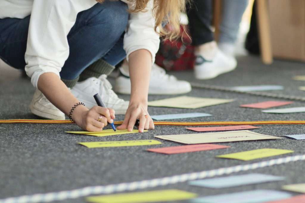 Person schreibt auf farbige Karten, die auf dem Boden ausgelegt sind. Weitere Karten und Teilnehmende befinden sich im Hintergrund eines Workshop-Raums. Das Bild zeigt eine praktische Gruppenübung im Rahmen eines Training Psychologische Sicherheit und verdeutlicht gemeinsames Arbeiten, Austausch und aktives Lernen.